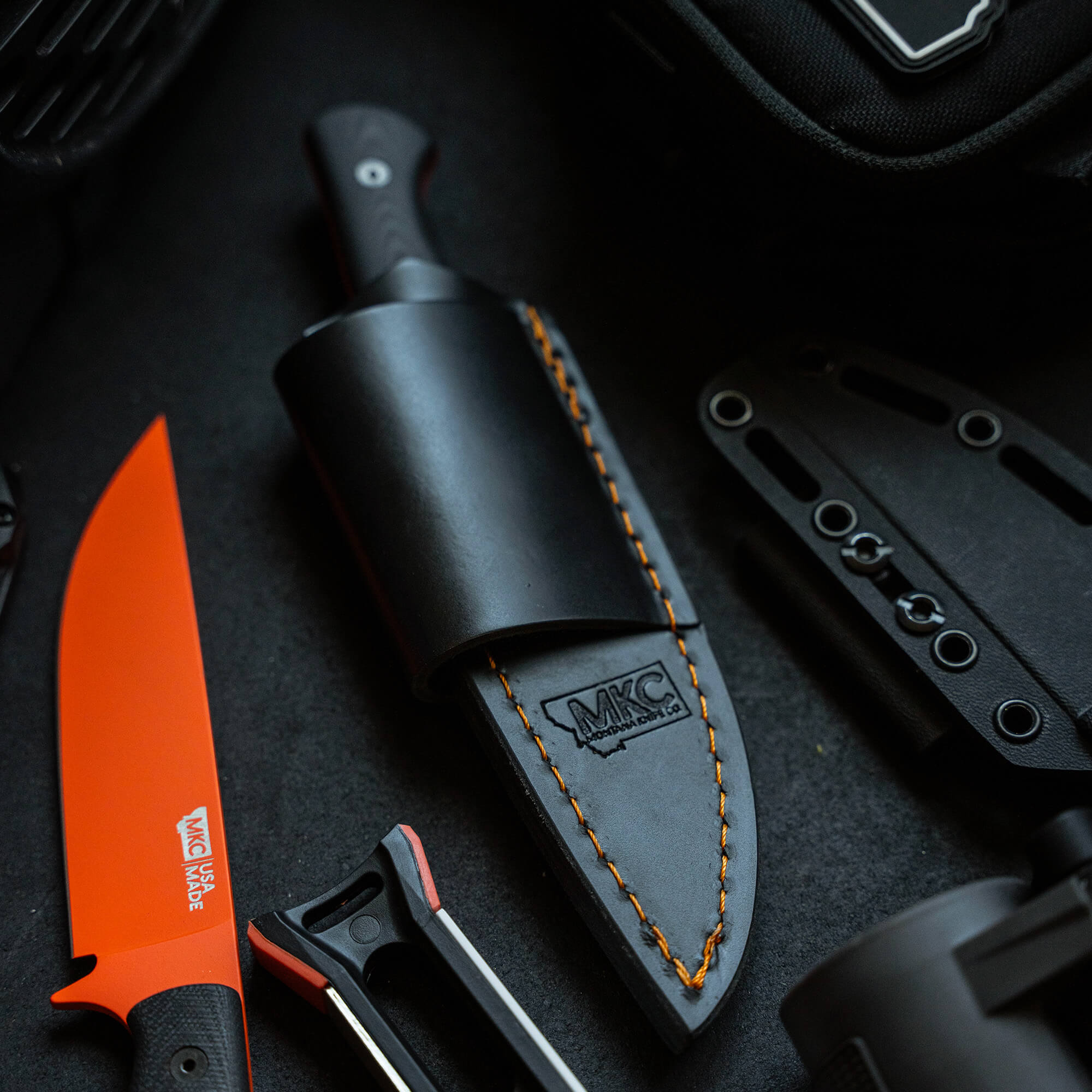 Black and orange Blackfoot 2.0 knife with a leather sheath for horizontal carry, displayed alongside tactical gear on a dark background.