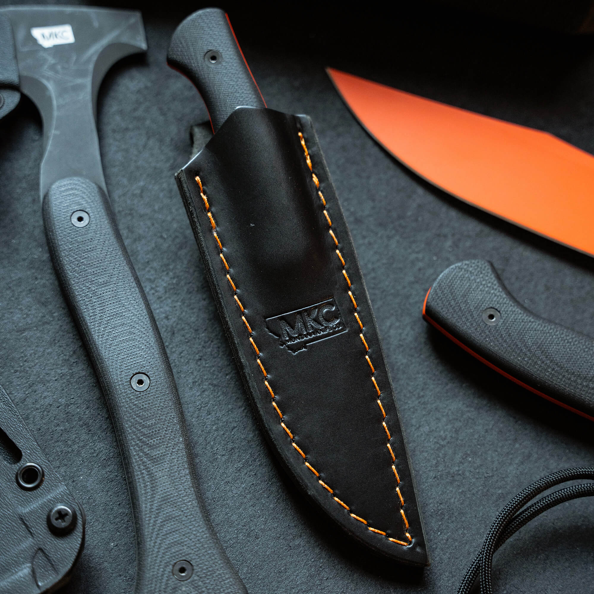 Alt text: "Close-up view of a black leather sheath with orange stitching and embossed MKC logo, designed for vertical carry, alongside a collection of black and orange handled knives and an axe, highlighting quality and craftsmanship."