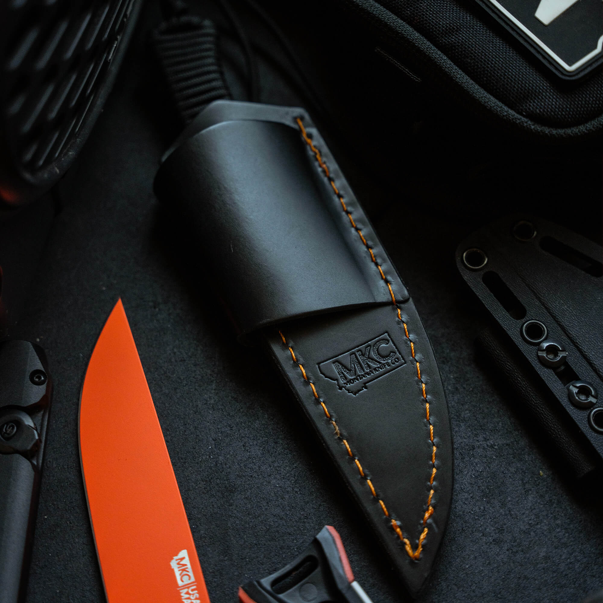 Black leather sheath with orange stitching, designed for horizontal belt carry, displayed beside a bright orange knife blade and various tactical gear.