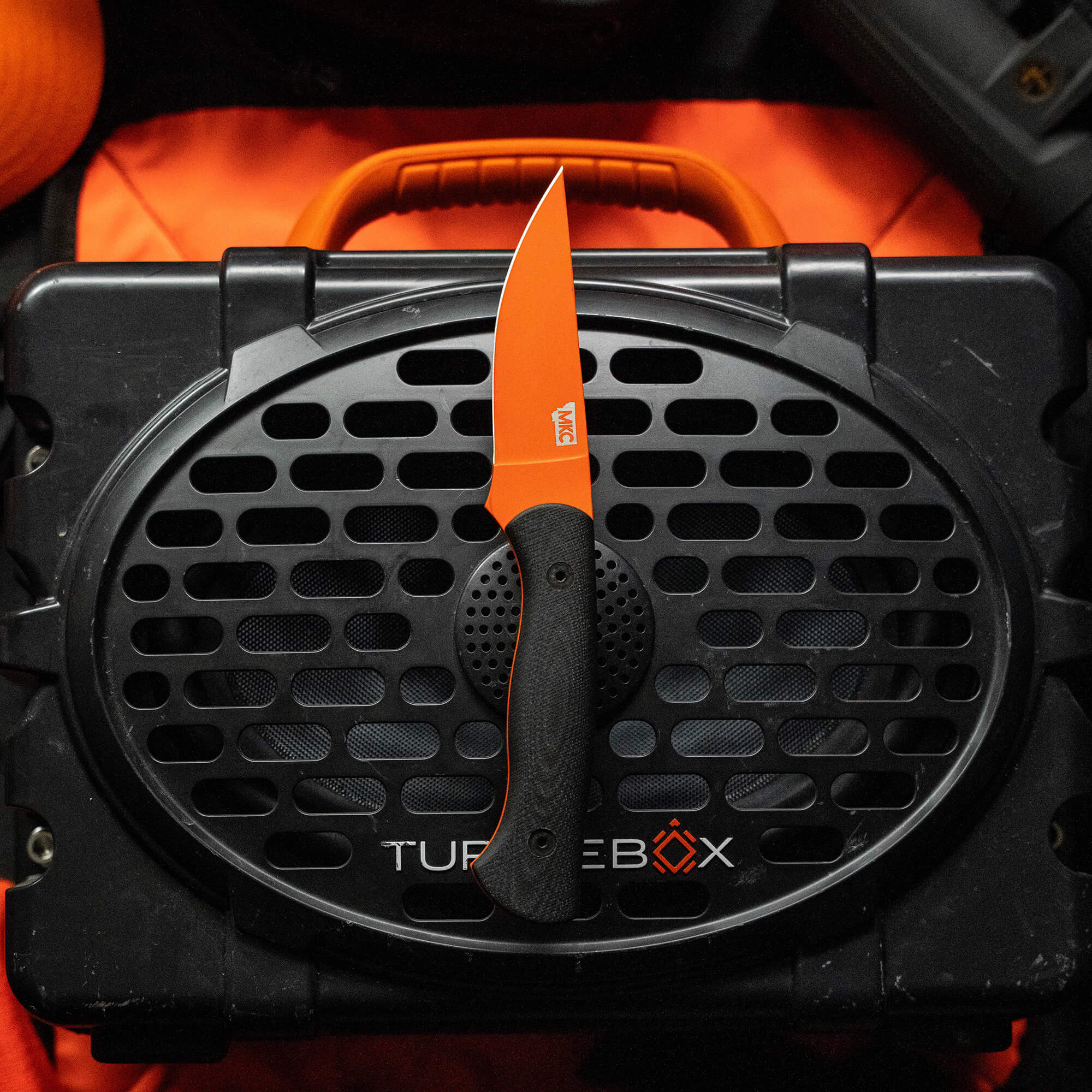 Alt text: "Blackfoot 2.0 fixed blade knife with blaze orange Cerakote finish on the blade, resting on a black TUFBX portable speaker grill with an orange carrying case visible in the background."