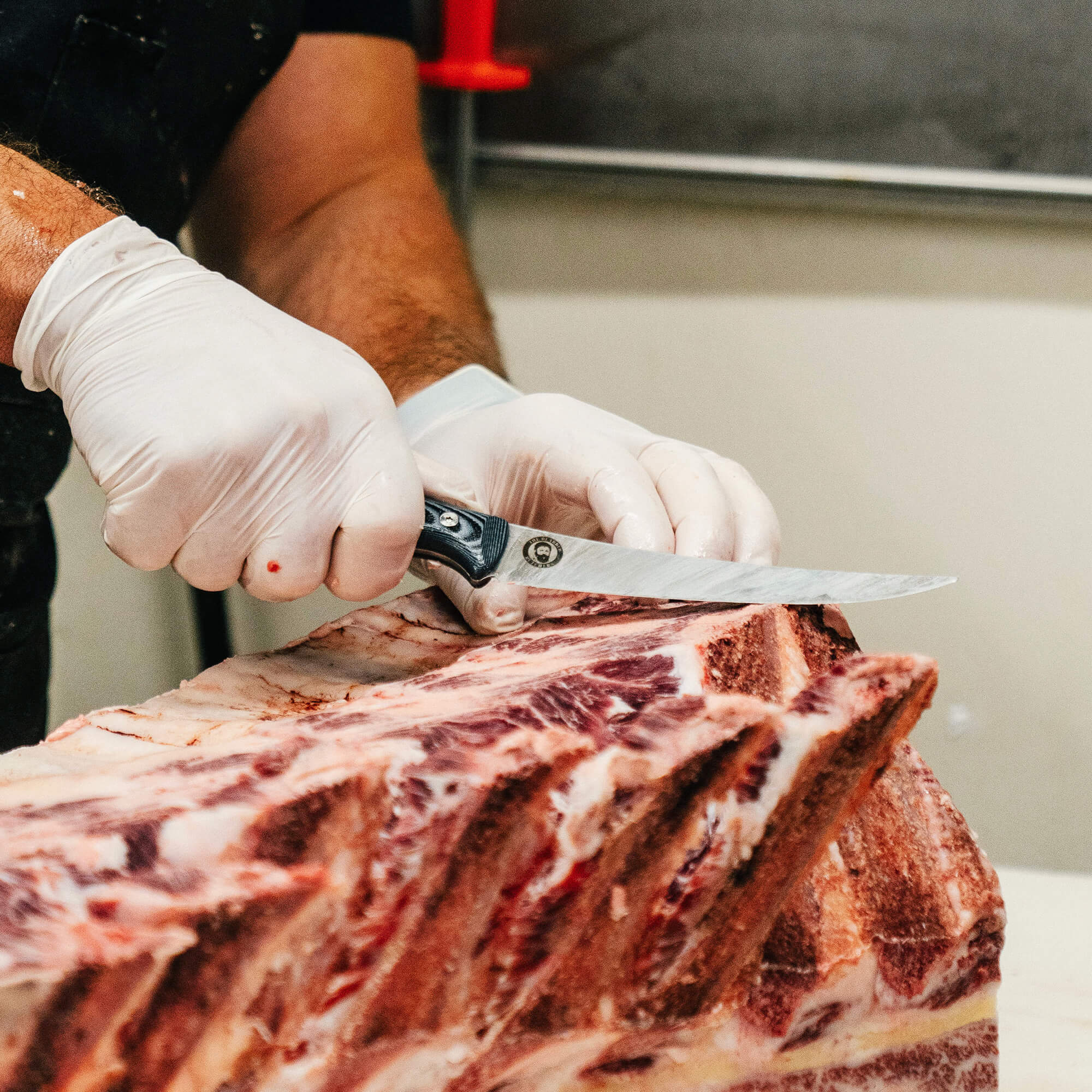 Alt text: "Close-up image of a Bearded Butchers brand boning butcher knife being used skillfully by a butcher in gloves to cut through large ribs of beef, highlighting the knife's sharp blade and ergonomic handle."