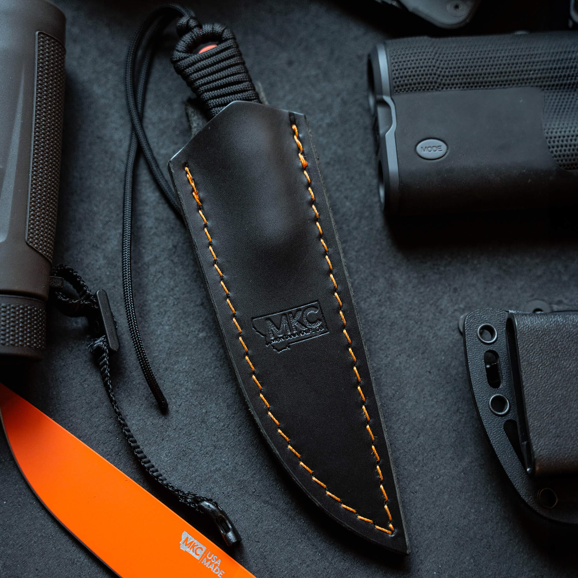 Alt text: Black leather sheath with orange stitching labeled "MKC", designed for vertical belt carry, surrounded by tactical gear including a flashlight and a partial view of an orange-handled knife.