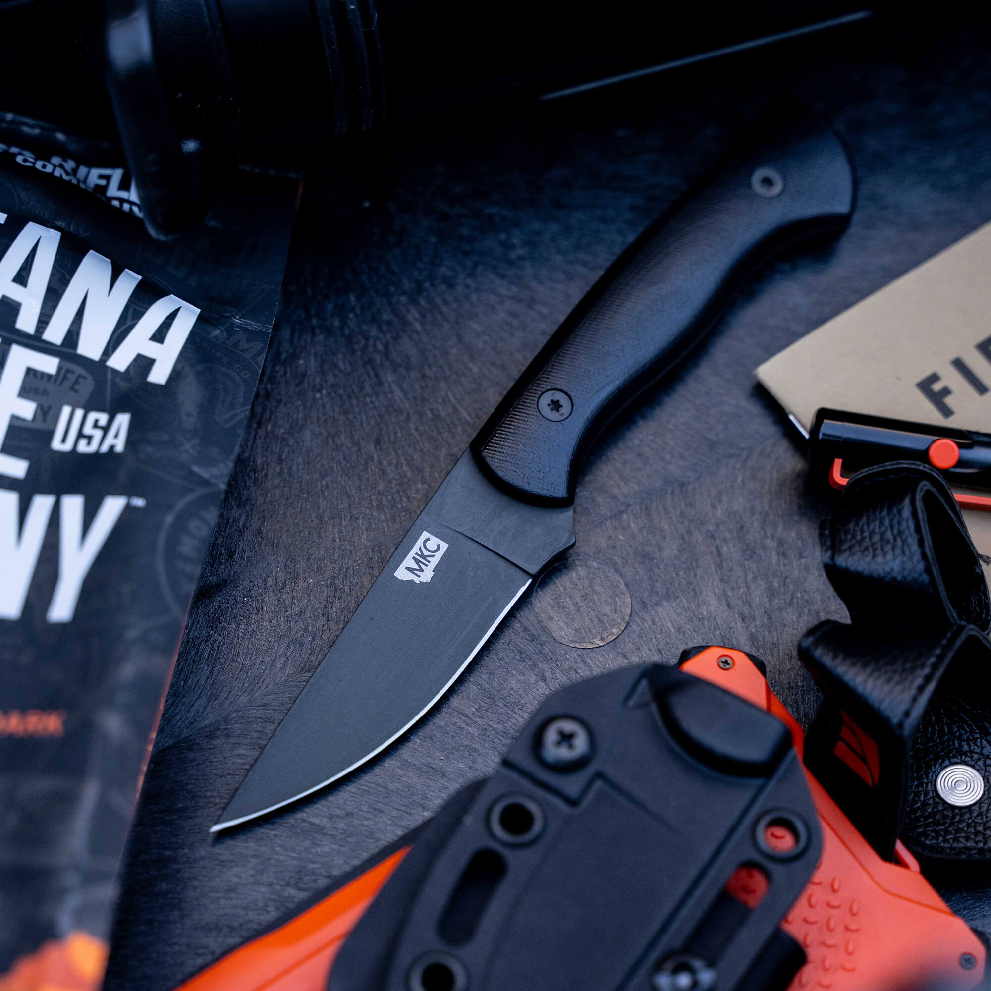 Alt text: "Blackfoot 2.0 tactical knife with a black handle and blade, displayed next to an orange and black holster, a headlamp, and outdoor survival gear including a topographic map."