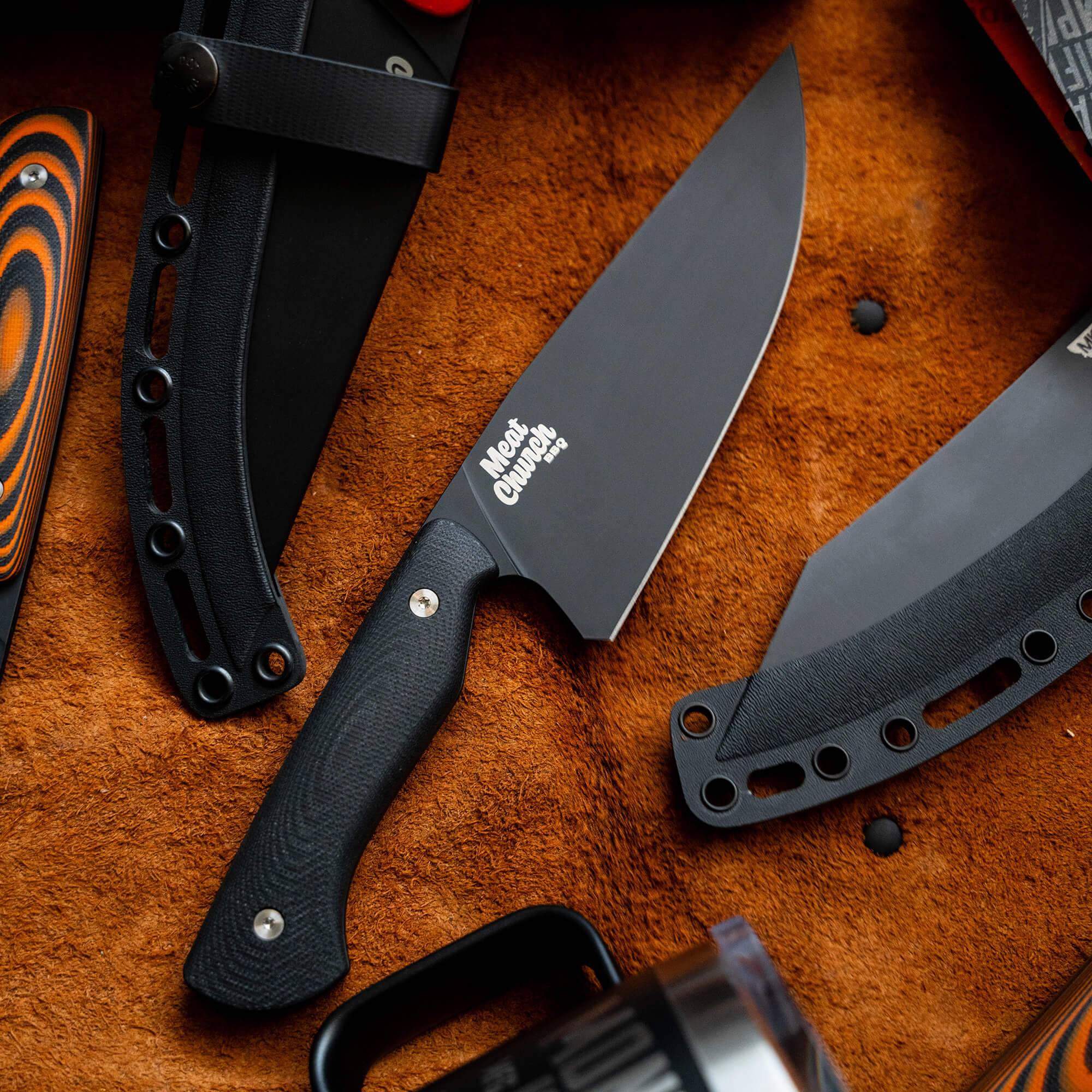 Alt text: "Close-up view of a Meat Church chef knife with a black handle and logo displayed prominently on the blade, alongside a black carrying sheath, set on a textured orange background."