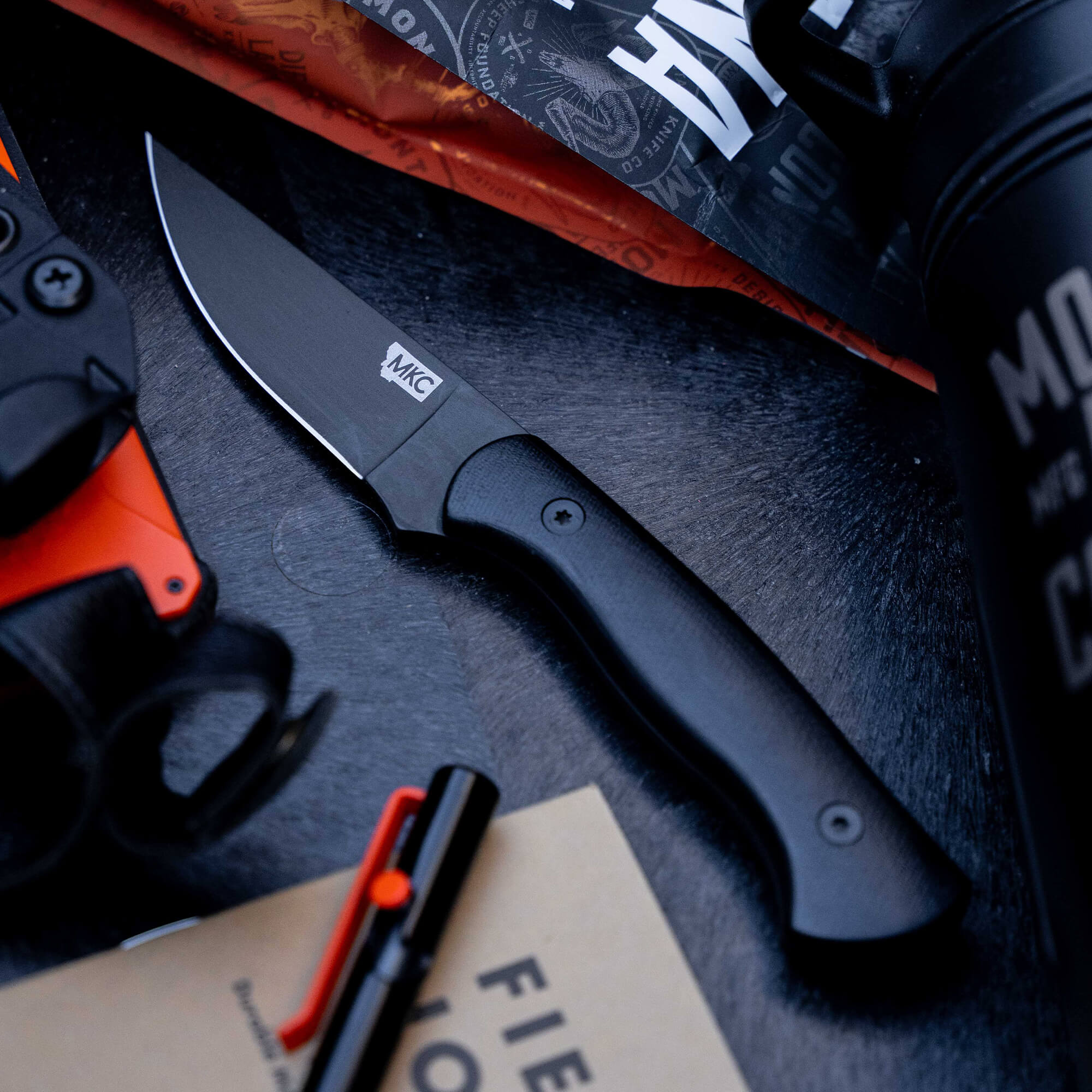 Alt text: "Close-up view of the Blackfoot 2.0 knife in black with a matte finish and branded handle, displayed alongside outdoor gear and survival tools on a dark textured surface."