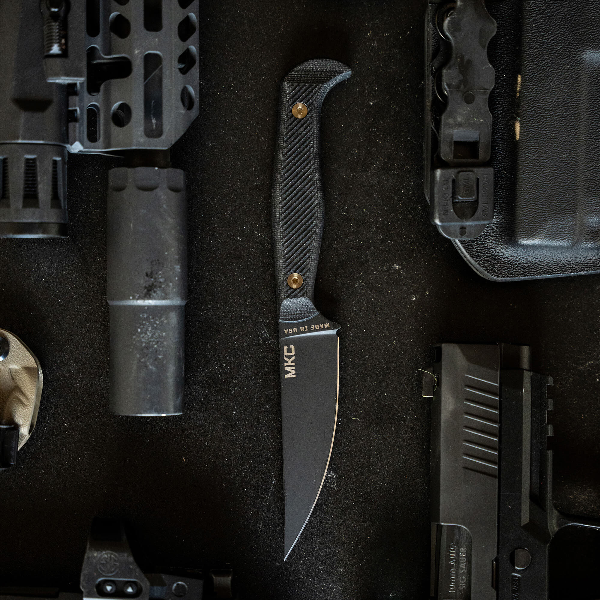 Black tactical knife, labeled MKC, with a curved handle and sharp blade, displayed amidst firearm parts and tactical gear on a dark, textured background.