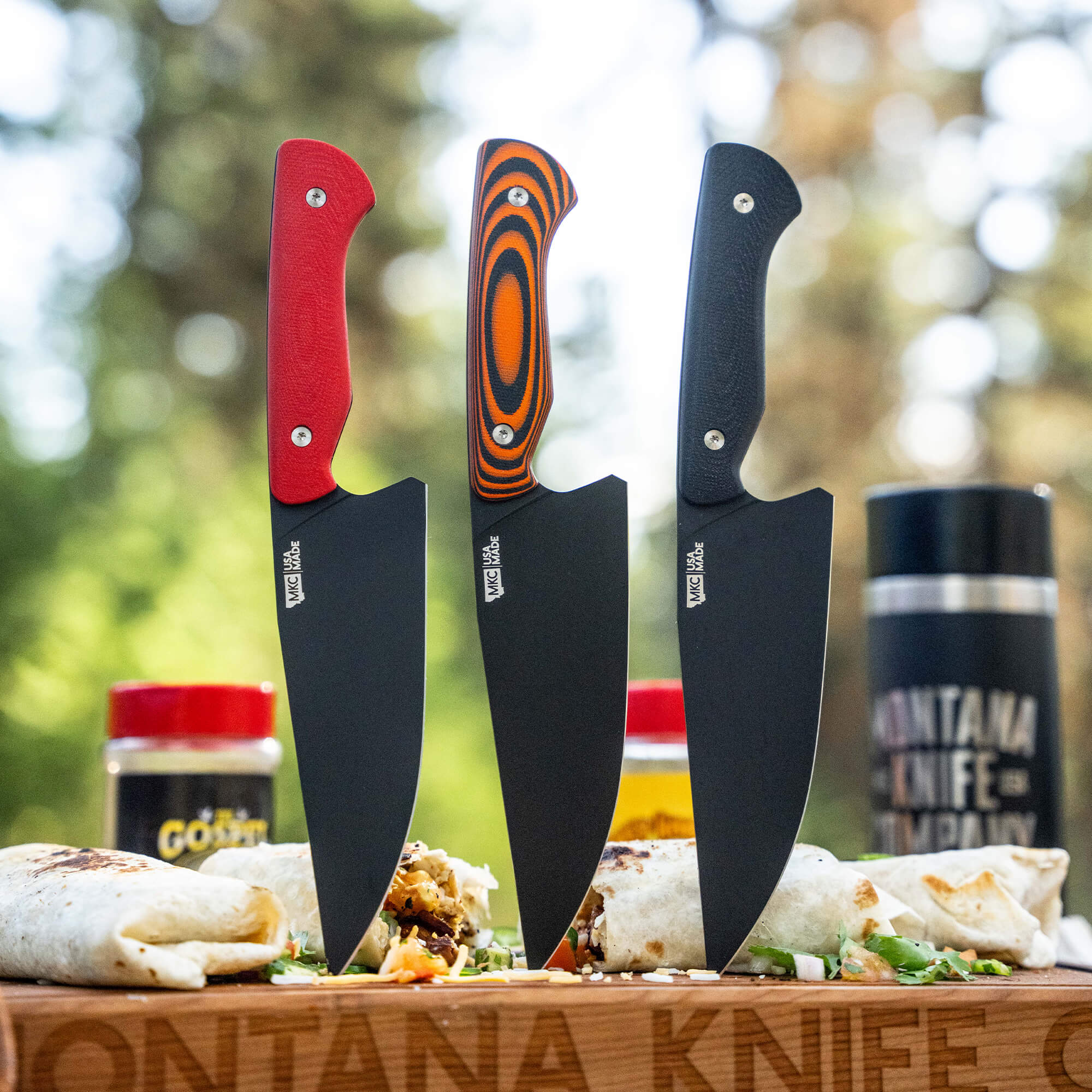 Three Meat Church chef knives with distinctive black blades and color-accented handles (red, multi-colored, and black) displayed outdoors on a cutting board with burritos, suggesting culinary use in a rustic setting.