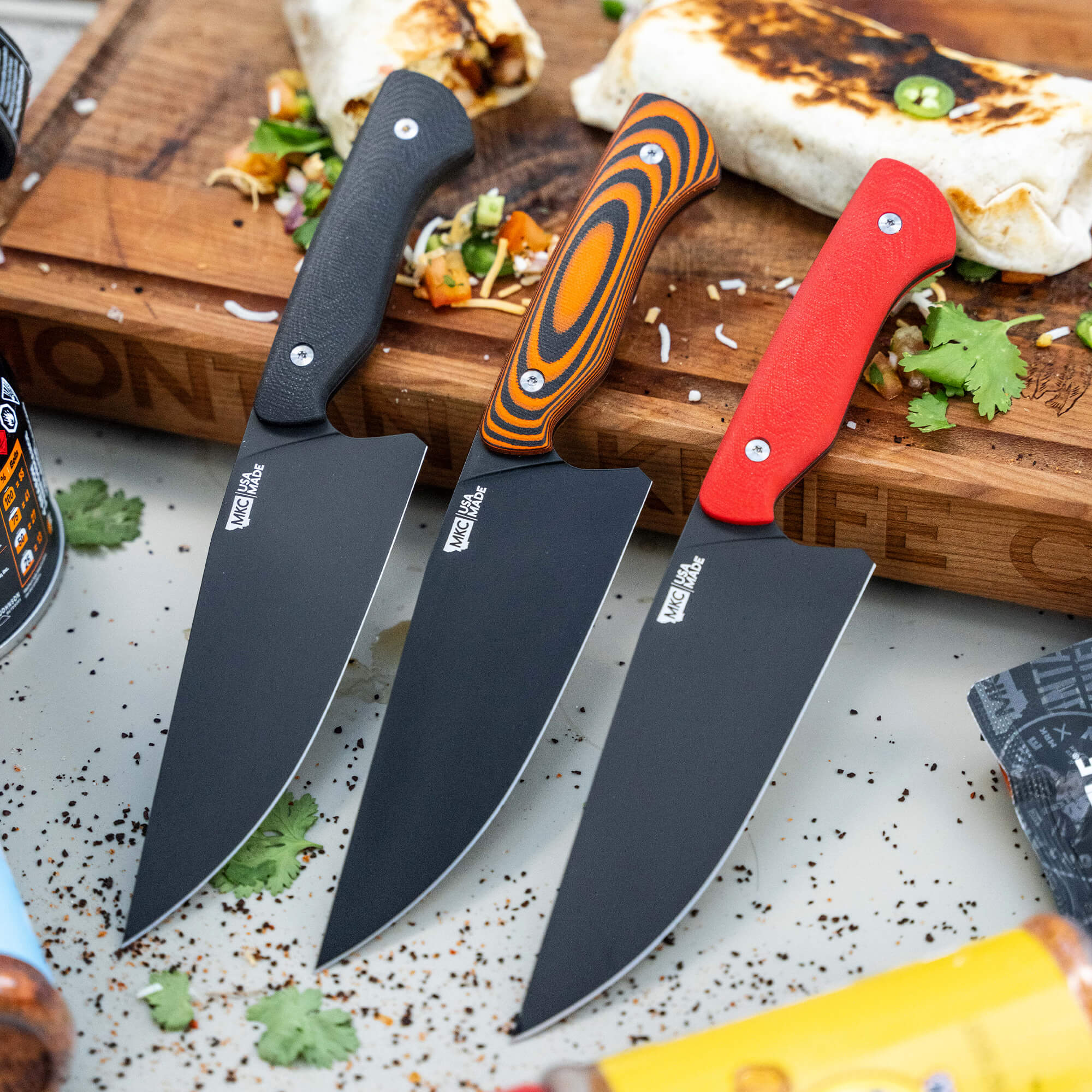 Alt text: Three Meat Church chef knives with stylish handles in black, red, and multicolored designs displayed on a wooden cutting board with chopped vegetables in the background, emphasizing sharp, modern culinary tools.