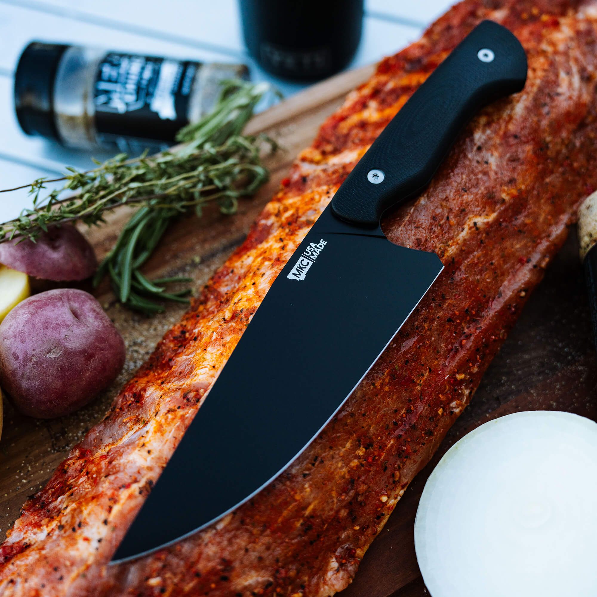 Alt text: "Meat Church chef knife with a black handle lying next to a seasoned rack of ribs on a wooden board, accompanied by fresh herbs, potatoes, and spices."