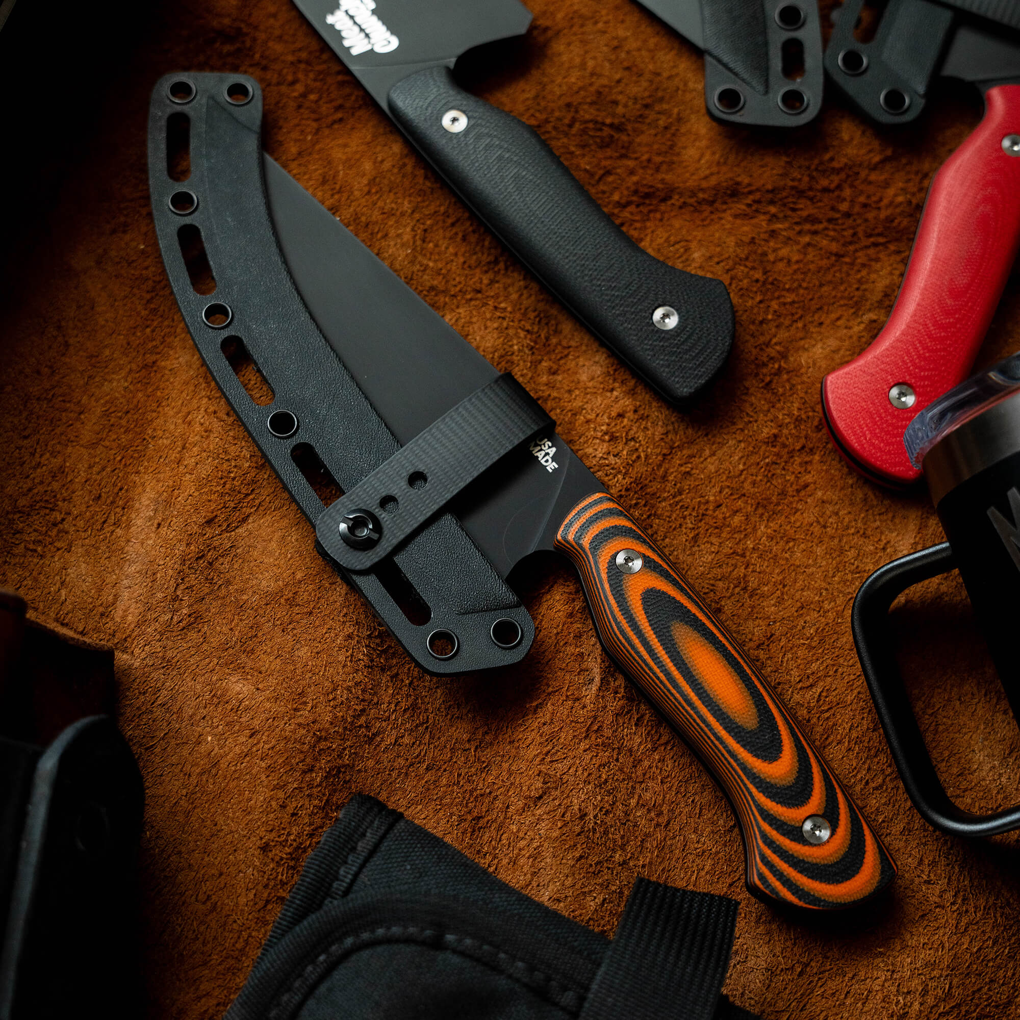 Alt text: Close-up of a Meat Church chef knife with a distinctive orange and black striped handle, displayed among various other knives, featuring a black protective sheath, on a brown textured background.