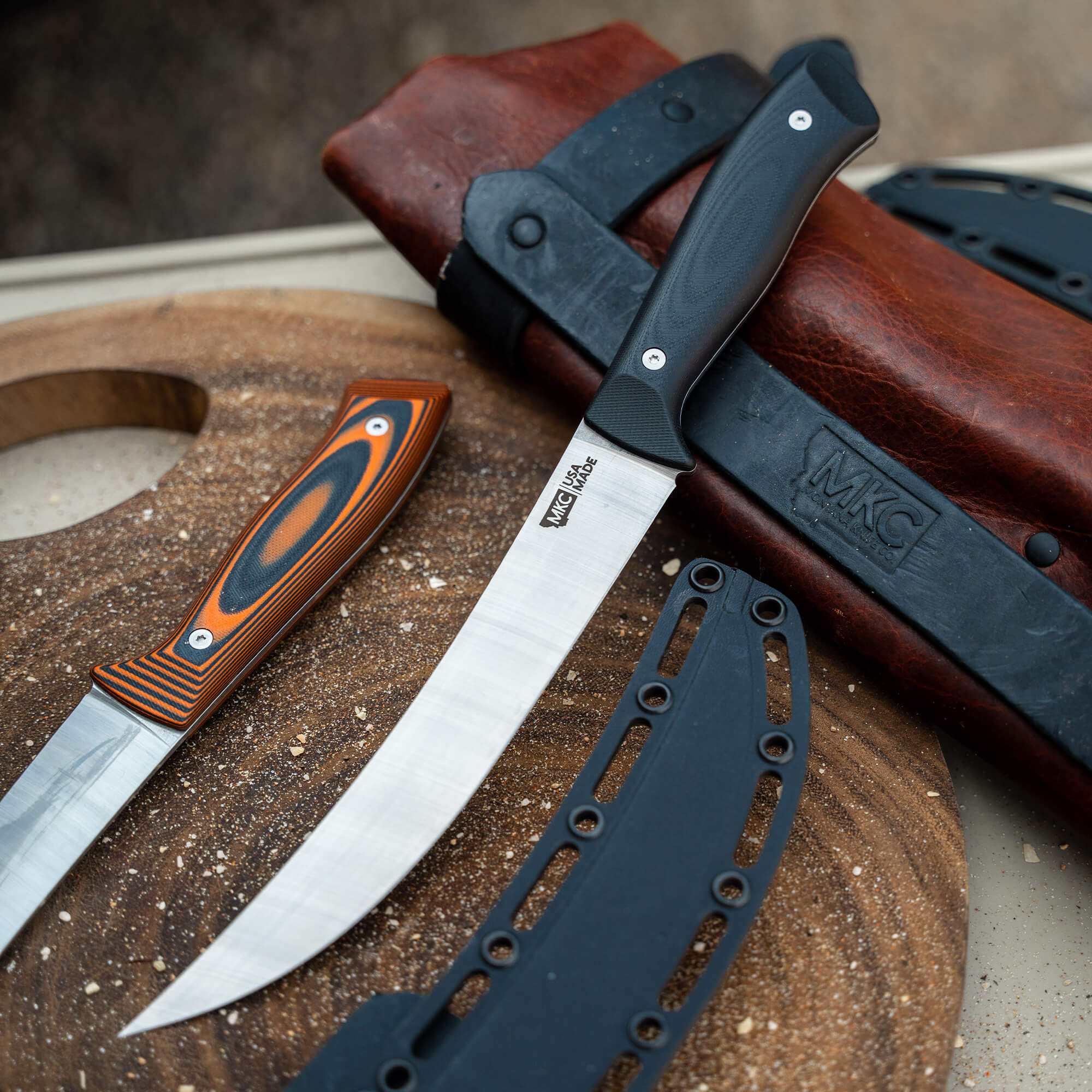 Alt text: "Bearded Butchers Breaking Butcher knives from a secret drop, featuring two robust utility knives with black and orange handles, displayed on a textured surface with leather sheaths and blade protector parts."