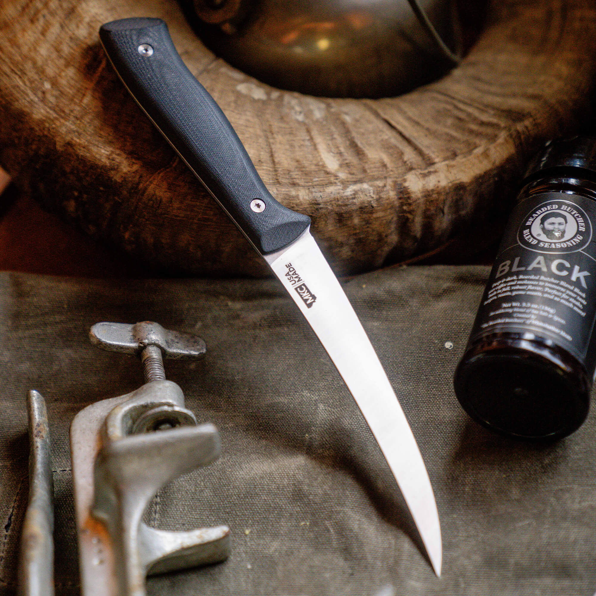 Alt text: "Bearded Butchers boning butcher knife placed on a rustic workbench, surrounded by tools and a bottle of seasoning, highlighting its sleek design with a black handle and stainless steel blade."