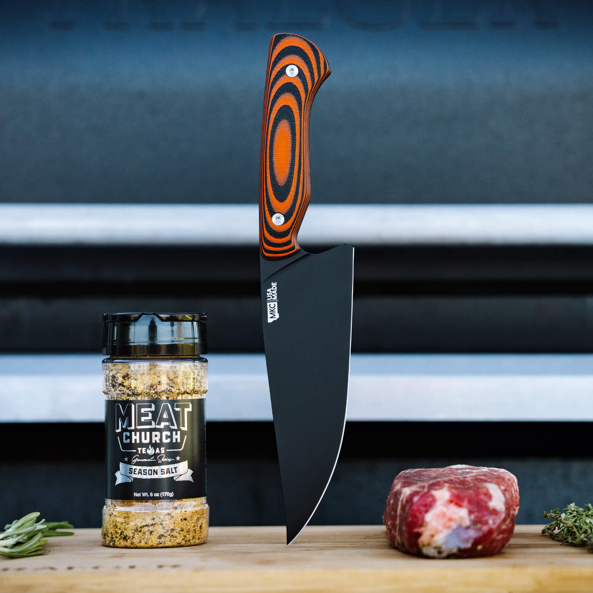 "Meat Church chef knife with a vibrant orange and black handle, displayed upright next to a jar of Meat Church Texas Season Salt and a piece of raw meat, set against a blurred outdoor grill background."