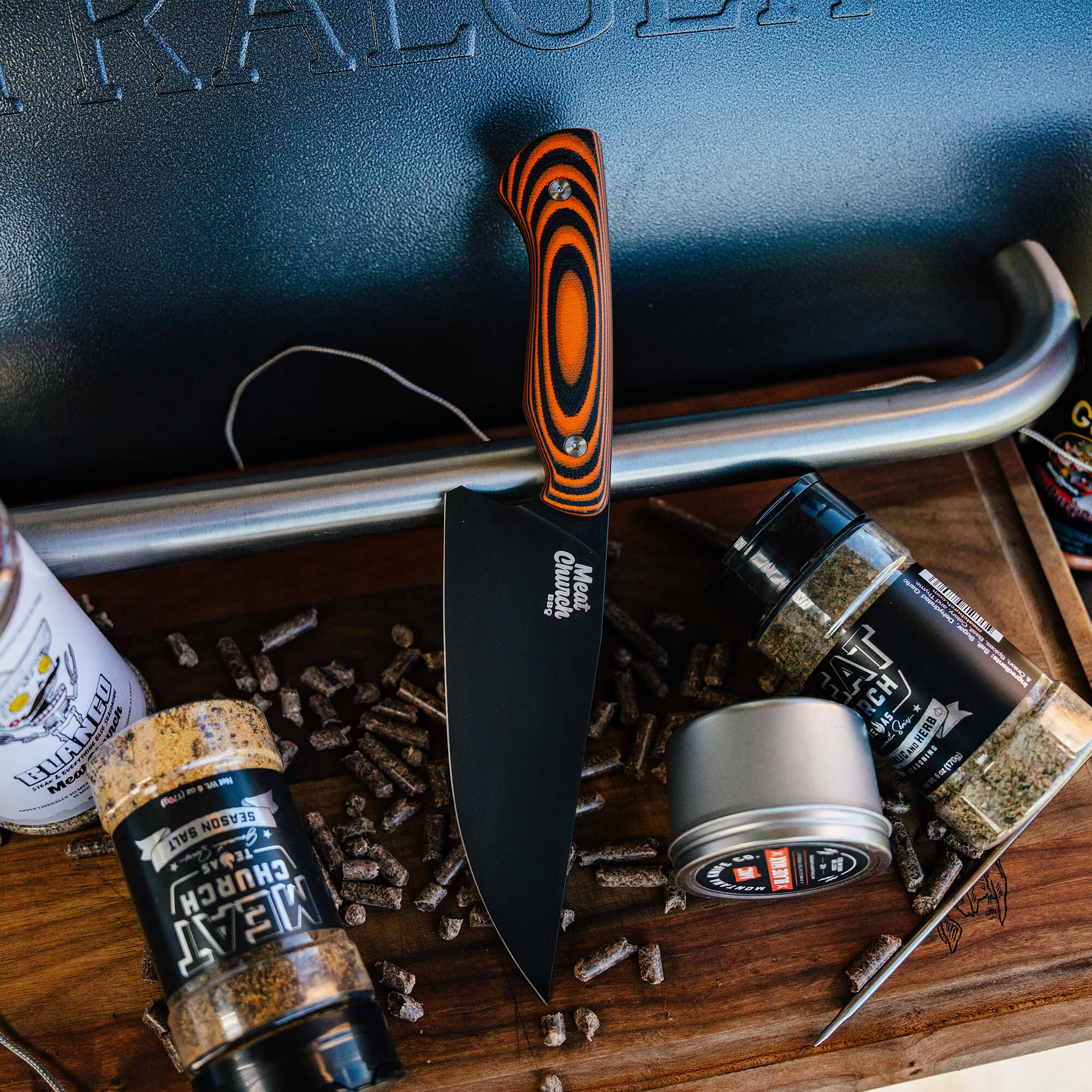 Alt text: "Meat Church chef knife with black blade and orange-black striped handle, displayed alongside BBQ rubs and pellets on a wooden surface."