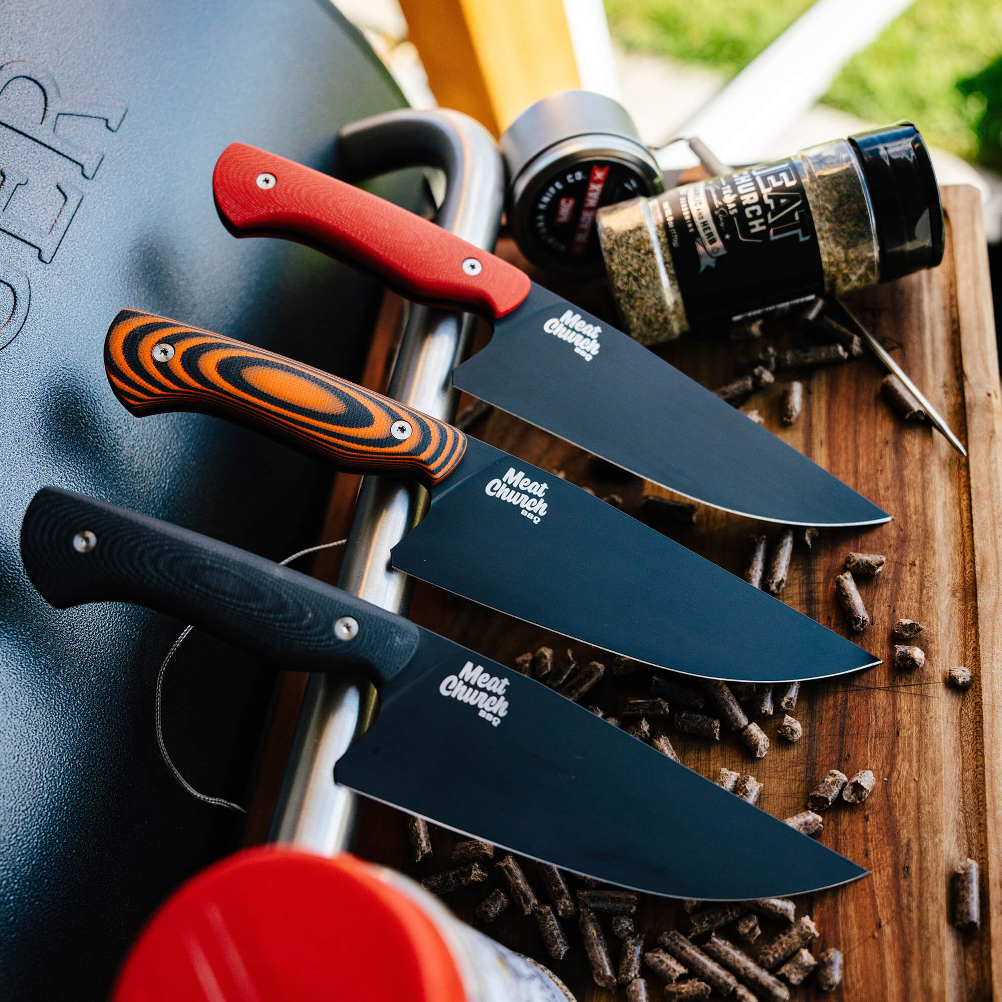 Alt text: "A vibrant display of Meat Church chef knives with assorted handle designs resting on a wooden board near a grill and spices. The knives feature black blades with the Meat Church logo, emphasizing their exclusive and stylish design."